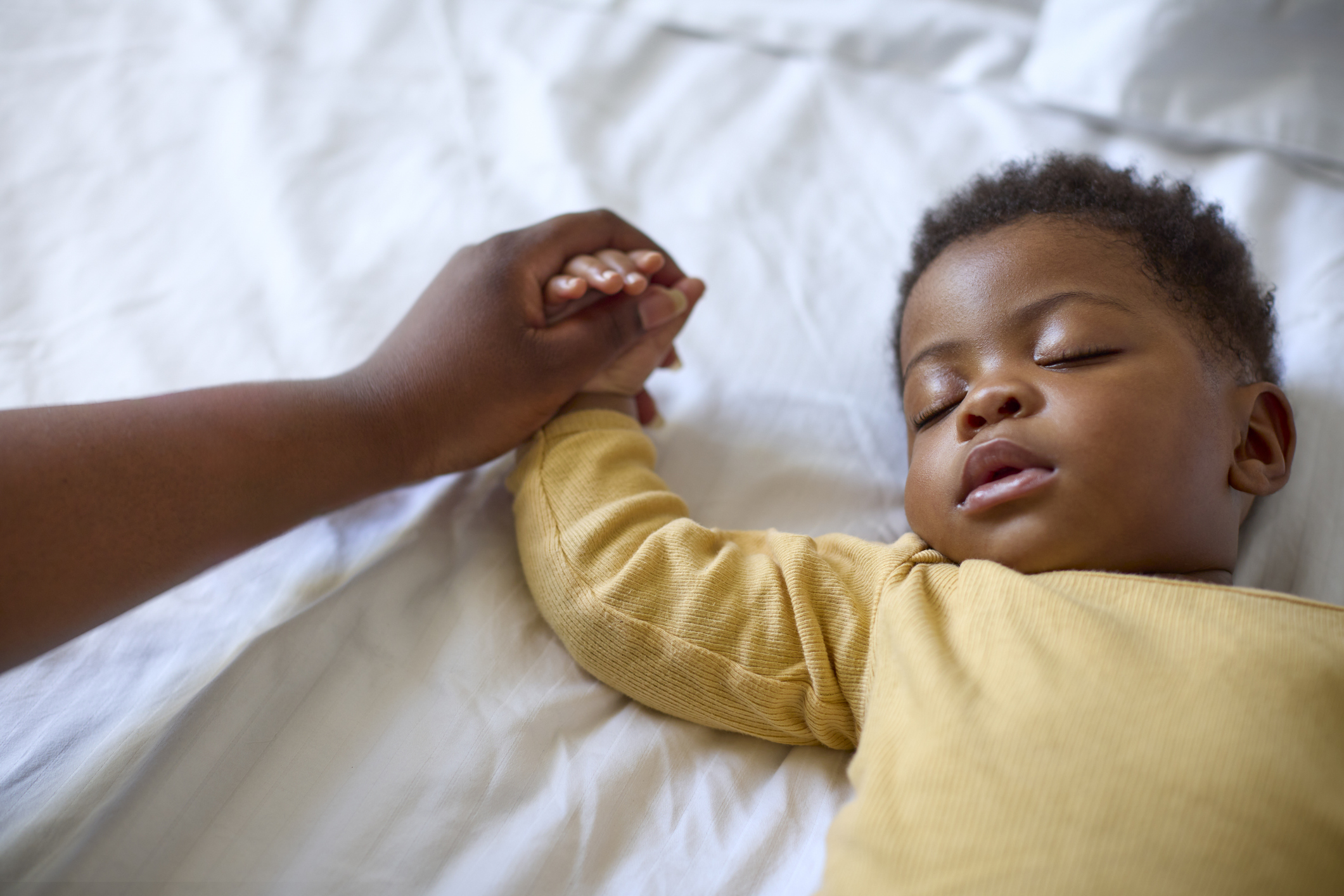Close Up Of Loving Mother Holding Baby Daughter's Hand As She Sleeps On Bed At Home